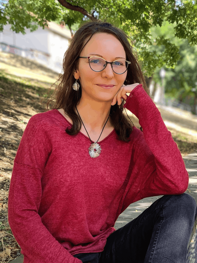 Frau mi langen braunen Locken, Brille und roem Pulli sitzt da und lächelt.